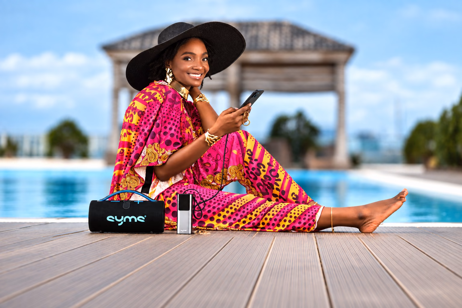 Simi sitting by a pool with a SYMS Wave speaker and a SYMS House Party power bank